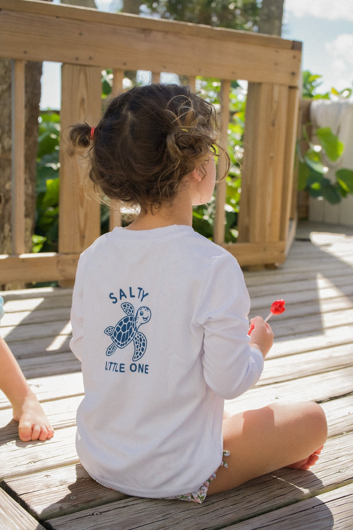 Back view of a child wearing the SRQutie Salty SPF long sleeve tee in white with sea turtle graphic, kneeling on a boardwalk
