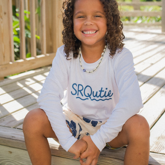 Child smiling while wearing the SRQutie Sun SPF long sleeve tee in white with SRQutie logo, sitting on a boardwalk