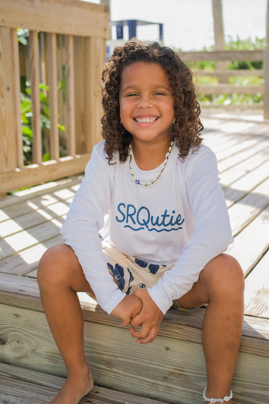Child smiling while wearing the SRQutie Sun SPF long sleeve tee in white with SRQutie logo, sitting on a boardwalk