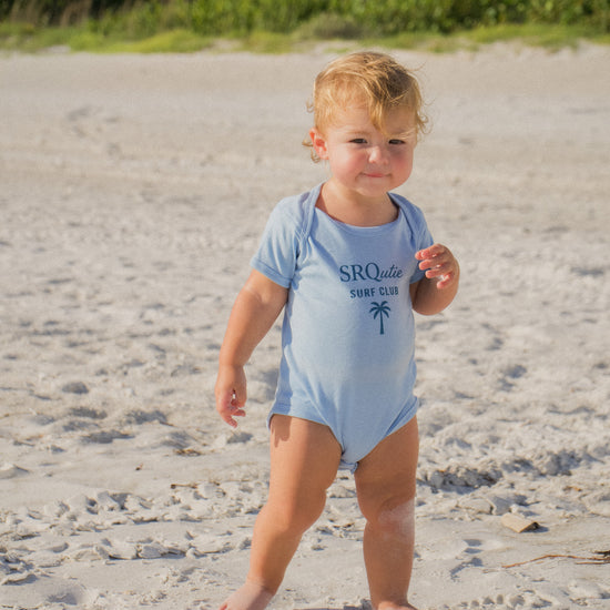 Baby wearing the SRQutie Surf Club onesie in light blue, standing on the beach