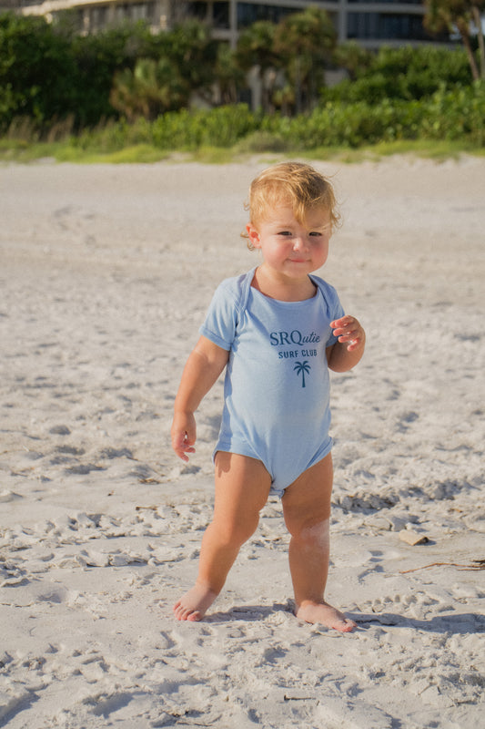 Baby wearing the SRQutie Surf Club onesie in light blue, standing on the beach