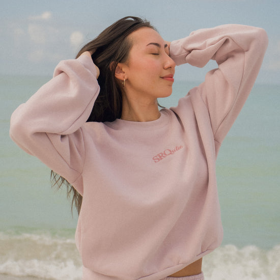 Woman on the beach wearing the SRQutie Logo Sweatshirt in Blush with the matching Logo Sweatpants