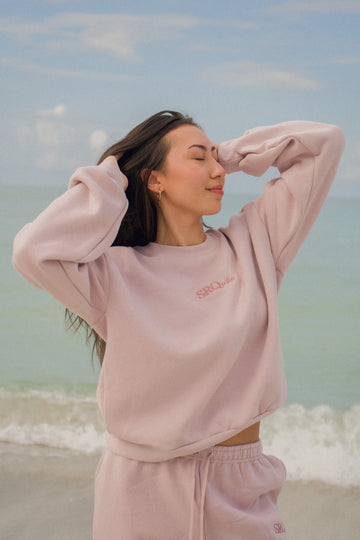 Woman on the beach wearing the SRQutie Logo Sweatshirt in Blush with the matching Logo Sweatpants